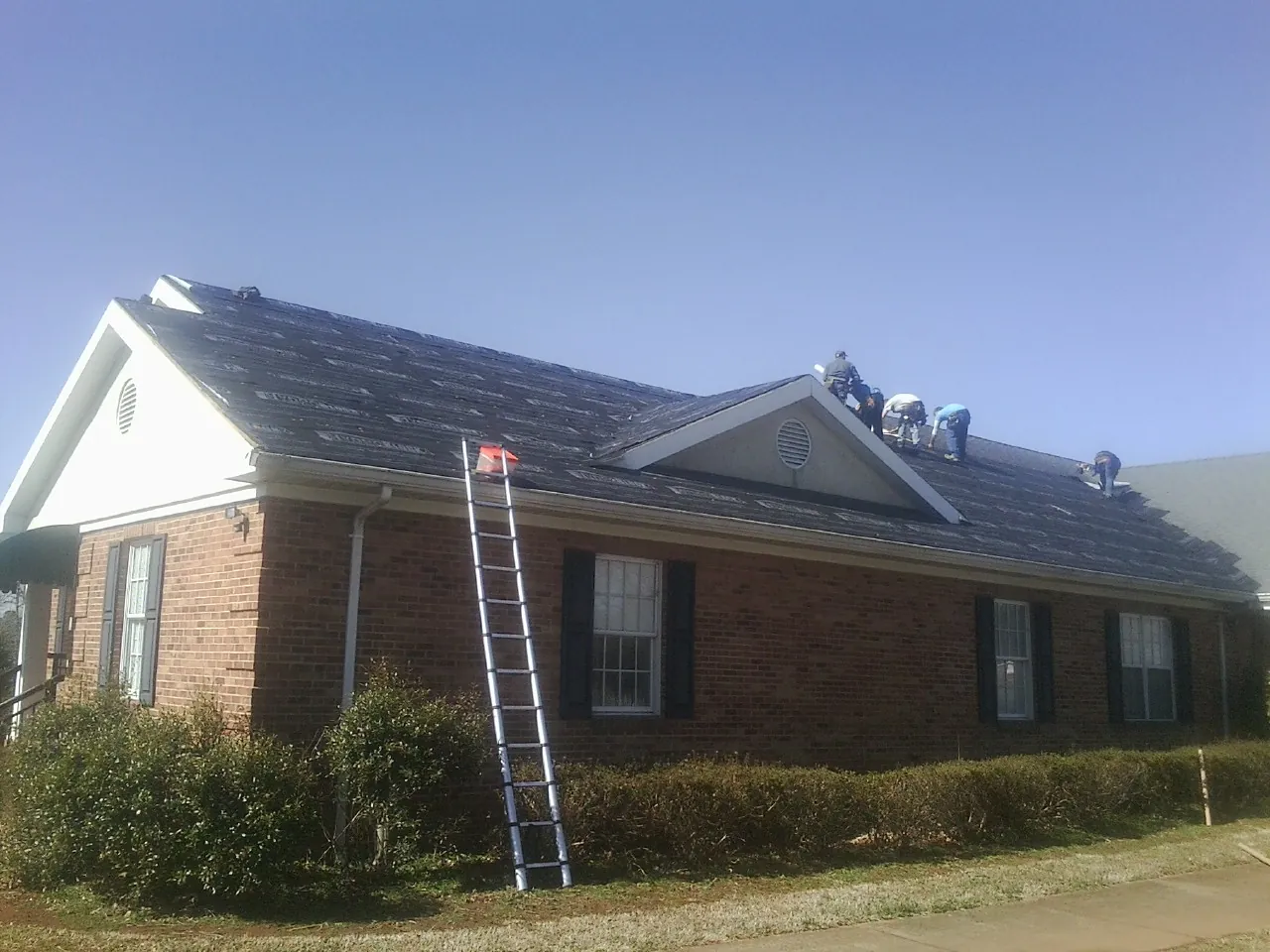 Crew tearing shingles off a brick ranch roof with a ladder set — Emergency Roof Repair work by our crew in Pursglove