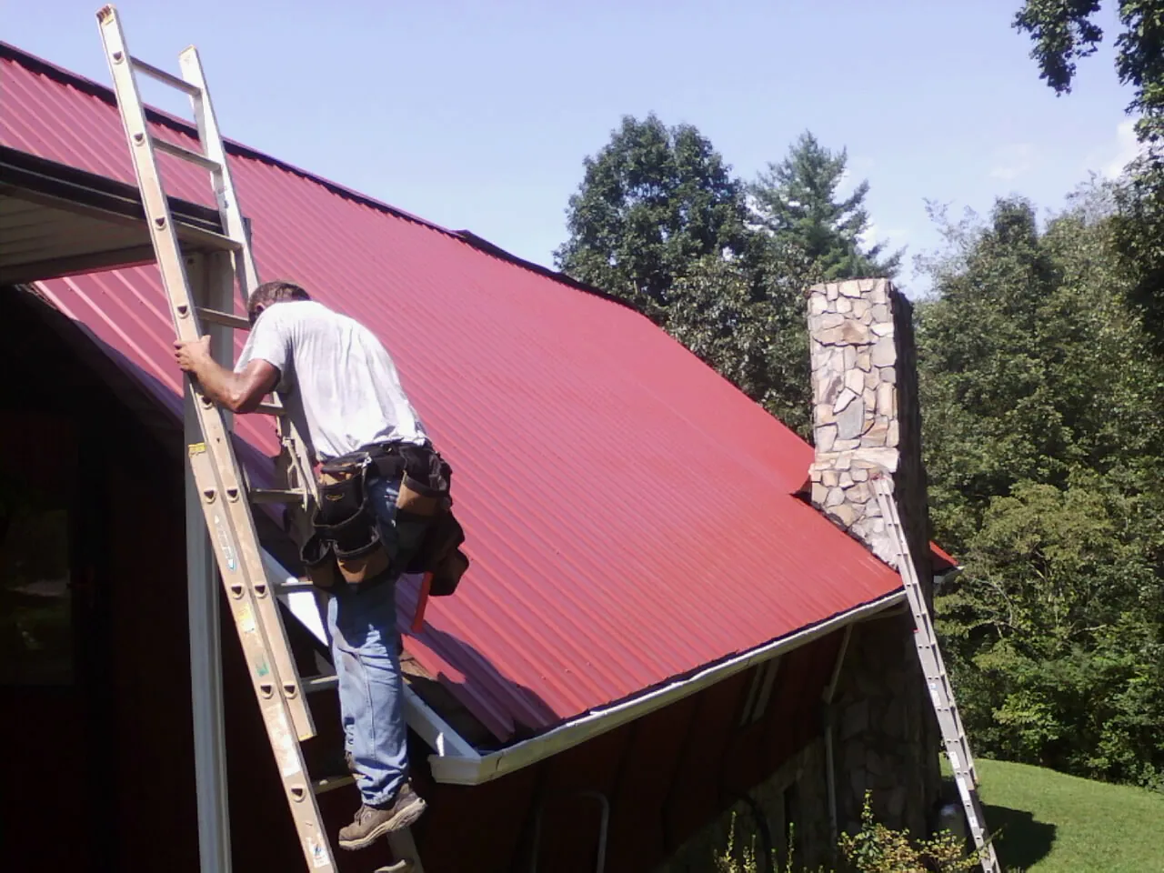 Roofer on a ladder beside a red metal roof and stone chimney — Soffit & Fascia Repair by Roofing Brothers in Pursglove